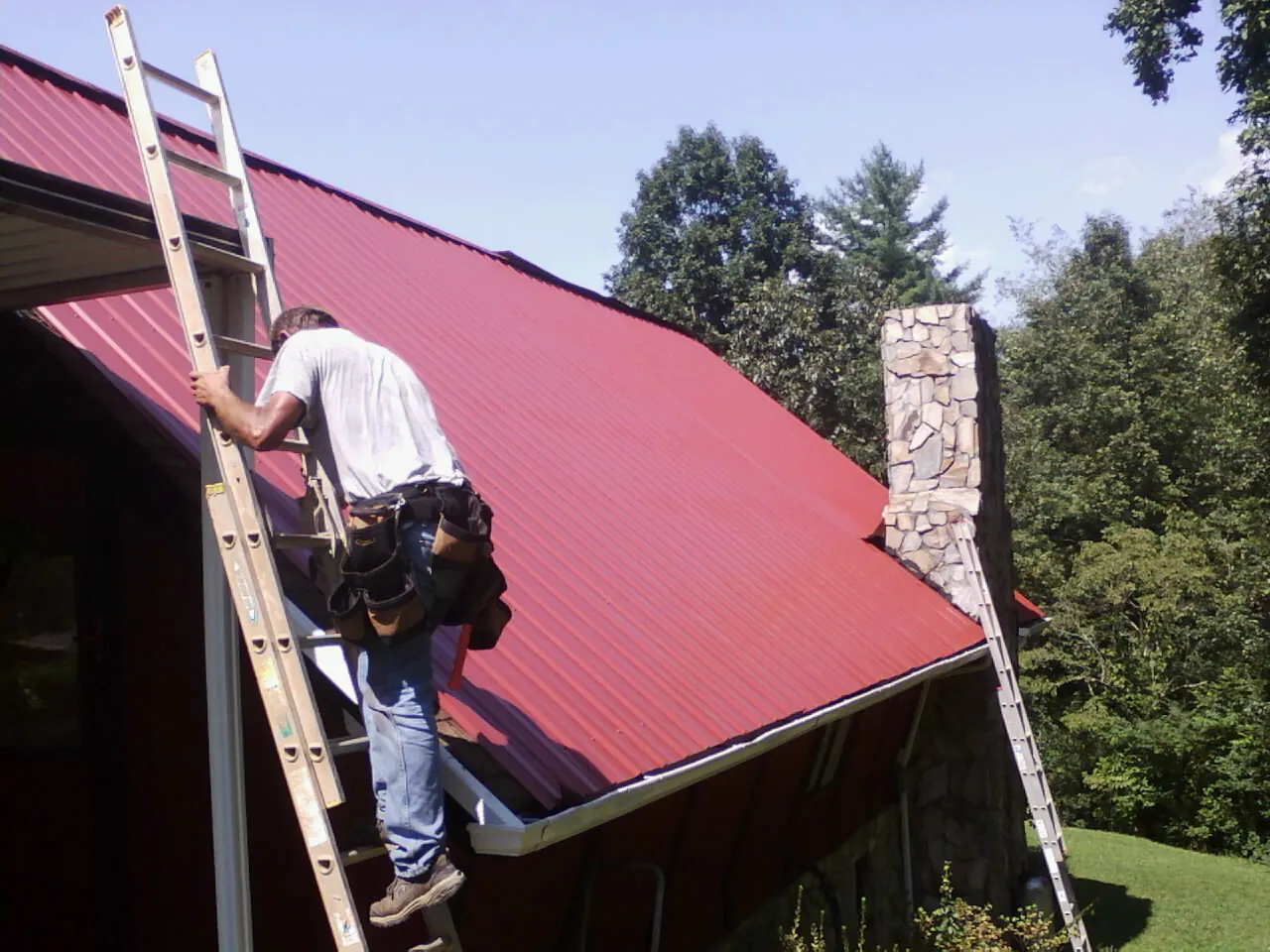 Roofer on a ladder beside a red metal roof and stone chimney — Soffit & Fascia Repair by Roofing Brothers in Gaylord